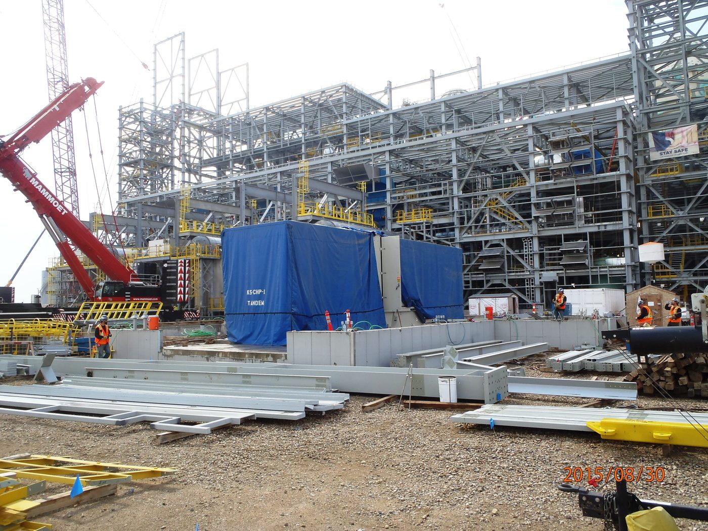 Heavy crawler crane setting modules against a large industrial steel structure under construction.