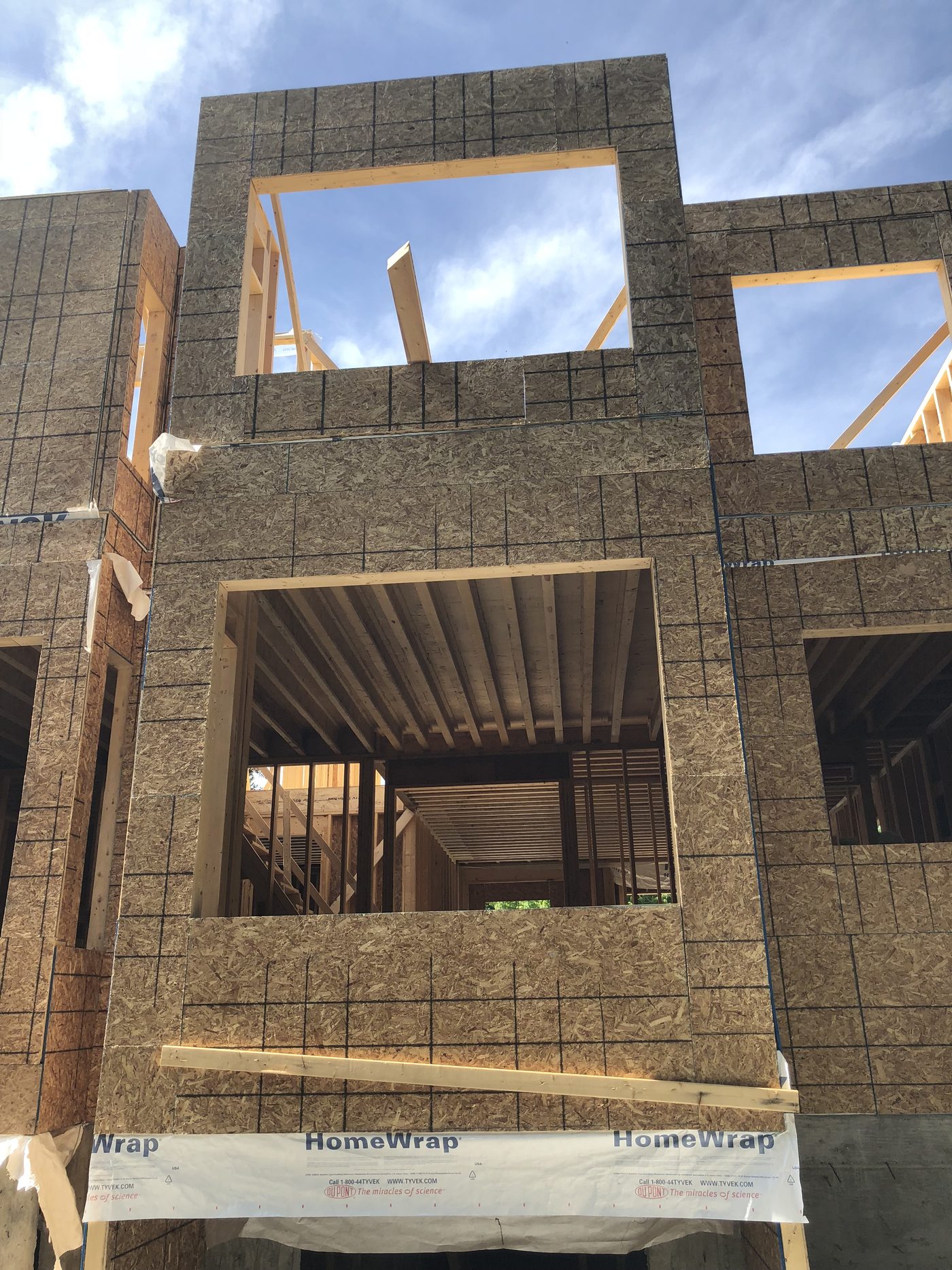 Wood-framed and sheathed house walls under construction, with engineered floor joists visible through a window opening.