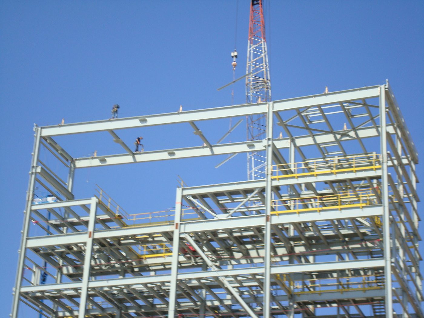 Multi-storey structural steel frame being erected by crawler crane against a clear sky, with ironworkers on the beams.
