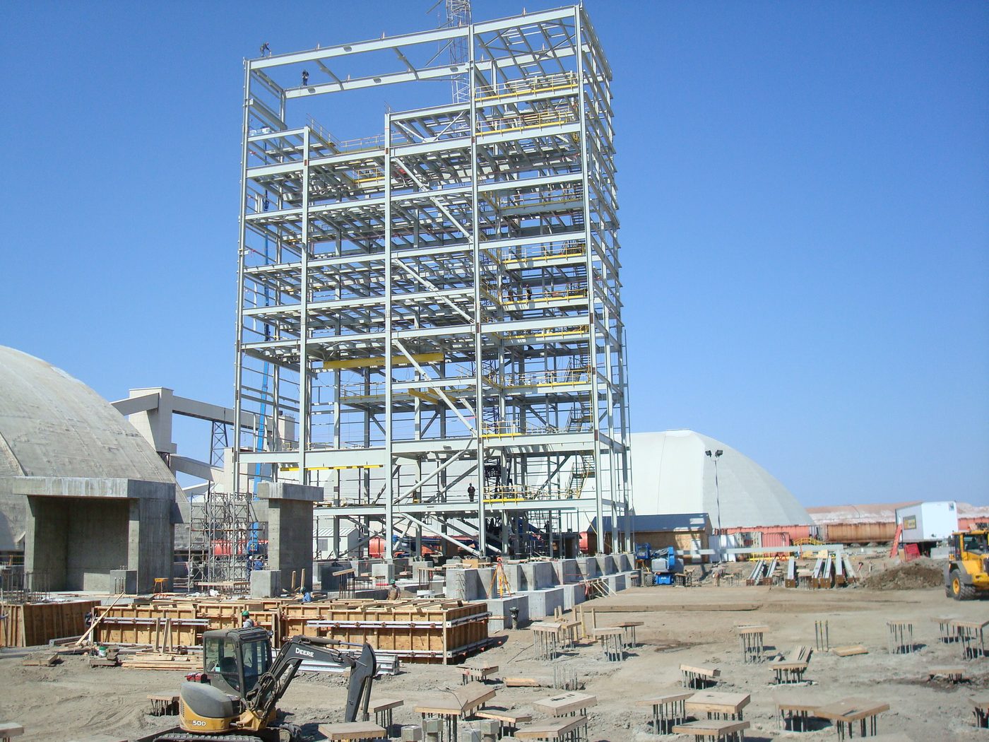 Tall multi-level steel plant structure under construction beside concrete storage domes, with foundations in the foreground.