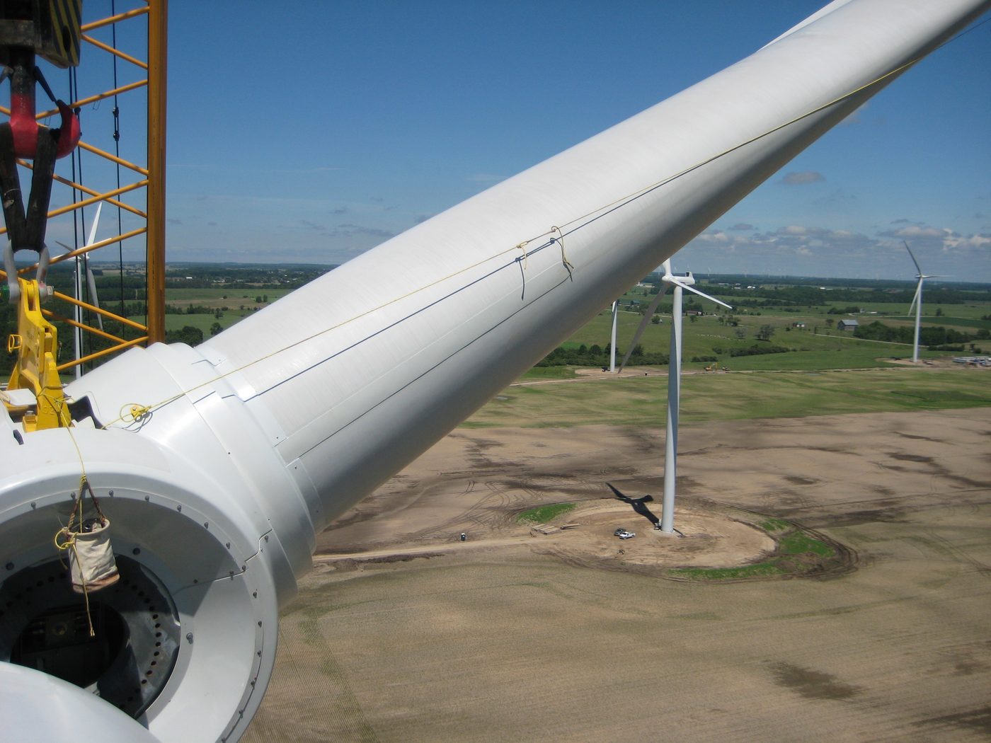 Turbine tower section suspended from a crane high above a wind farm and surrounding farmland.